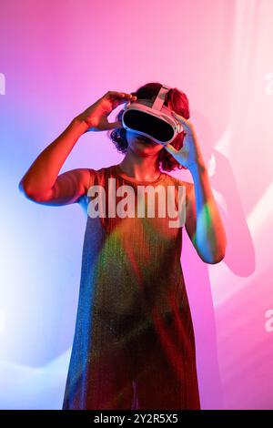 A young woman in a shimmering dress experiences a virtual world using a white VR headset, highlighted by vibrant pink and blue lights. Stock Photo