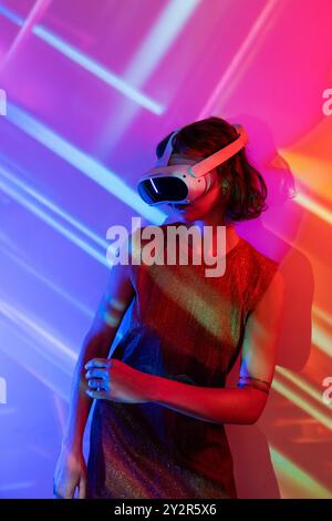 A young woman explores a vibrant virtual world using a sophisticated VR headset. Colorful lights and abstract projections enhance her futuristic exper Stock Photo