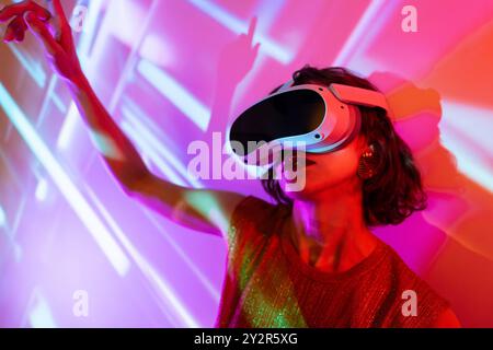 A young woman in vibrant lighting wearing a virtual reality headset, exploring the digital world of the metaverse. Stock Photo