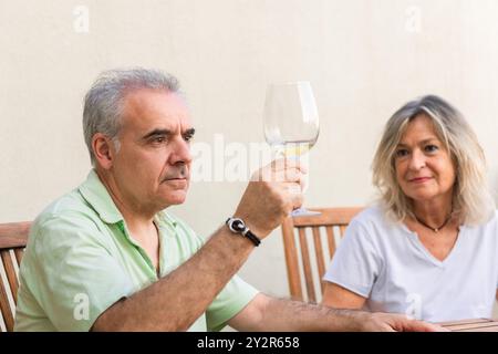 A mature man and woman savor a glass of white wine together on a wooden patio, depicted in a candid, casual setting with a serene backdrop Stock Photo