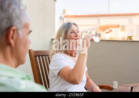 A mature woman sips wine while enjoying time on a balcony with her partner, showcasing a moment of leisure and connection The background features a ca Stock Photo
