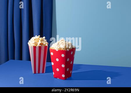 Two popcorn containers, one red with white stripes and the other red with white polka dots, sit on a blue table against a coordinating blue curtain ba Stock Photo