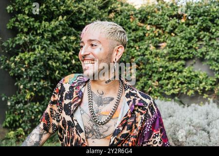 Colorful portrait of a joyful young man with vibrant hair and detailed tattoos, wearing a bold patterned shirt, laughing in a lush garden environment Stock Photo