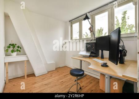 A modern, well-lit home office featuring dual computer monitors, a sleek desk, and a comfortable rolling stool. Stock Photo