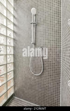 A sleek, contemporary shower head and hose set against a backdrop of small gray tiles and glass blocks on a bathroom wall. Stock Photo
