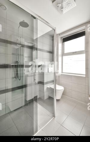 A sleek modern bathroom featuring a glass shower, white fixtures, and tiled walls bathed in natural light. Stock Photo