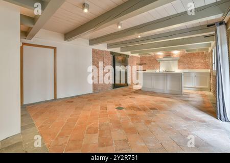 An empty rustic kitchen space featuring exposed brick walls, a white kitchen island, terracotta tile flooring, and wooden beams. Stock Photo