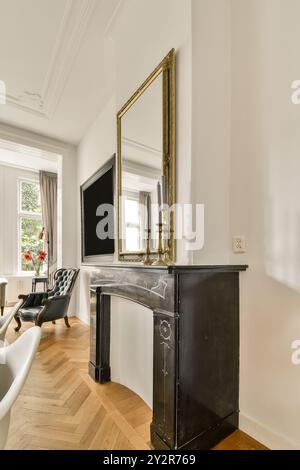 A refined living room featuring a black antique fireplace adorned with a large golden frame mirror and a cozy seating area by the window. Stock Photo
