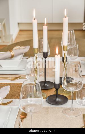 An inviting dinner table with lit white taper candles in stylish holders, surrounded by clear wine glasses, set against a neutral backdrop. Stock Photo