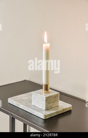 A tall, lit white candle set in a stylish marble holder placed on a dark wooden table against a neutral background. Stock Photo