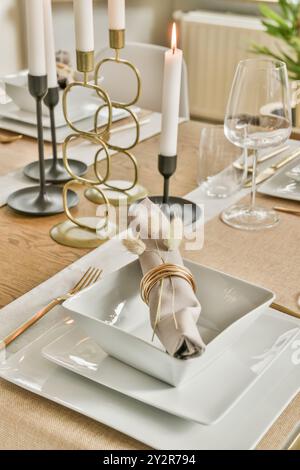 A contemporary table setting featuring white square plates, gold cutlery, and lit candles in stylish holders. Stock Photo