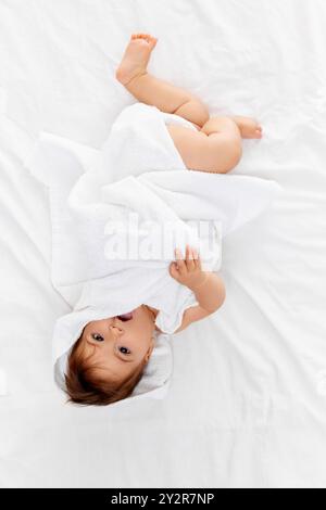 Wrapped in fluffy white towel, baby girl playfully peeks out, gazing curiously up at camera, her soft hair tousled from her playtime. Stock Photo