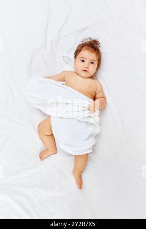 Wrapped in fluffy white towel, baby girl playfully peeks out, gazing curiously up at camera, her soft hair tousled from her playtime. Stock Photo