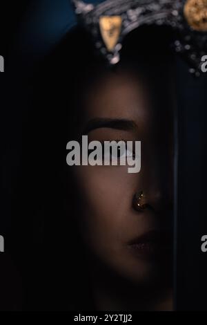 A close-up vertical portrait of Southeast Asian woman's shadowed face, with half obscured from view by an object Stock Photo