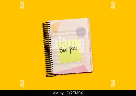 A spiral notepad with a sticky paper with See You short phrase handwritten on it on a yellow background Stock Photo
