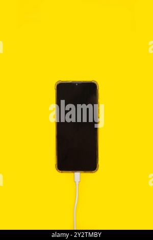 A smartphone connected to a charging cable on a yellow background Stock Photo