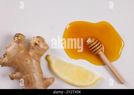A flat lay of fresh ginger root, a slice of lemon, and honey drizzled on a white surface. A small wooden honey dipper rests in the honey, creating a n Stock Photo