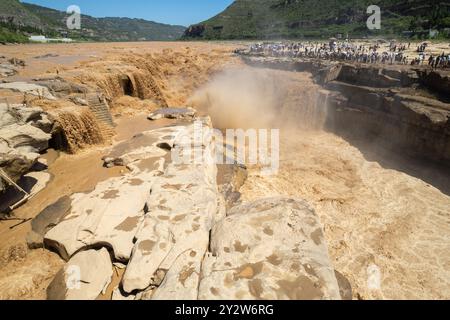 Tourists observing the powerful Hukou Waterfall on the Yellow River in China, surrounded by rocky terrain and lush green hills under a clear sky. Stock Photo