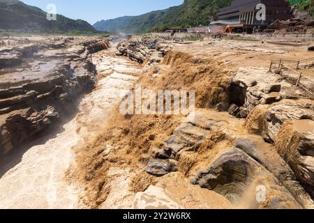 Hukou Waterfall on the Yellow River with muddy waters cascading down rocky terrain surrounded by mountainous landscape and tourists observing. Stock Photo