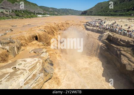 Tourists gather to view the majestic Hukou Waterfall on the Yellow River, China, surrounded by rocky cliffs and lush green hills. Stock Photo