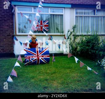 Young boy posing with home-made Union Flag sign for Queen Elizabeth II Silver Jubilee in June 1977 in the front garden of a home property with bunting ready for a street party Stock Photo