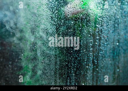 A glass window covered in water droplets and condensation, with soft green shapes blurred in the background, tropical vbe Stock Photo