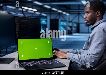 African american man working on laptop with green screen monitor in modern data center. IT expert doing IT infrastructure management on server racks in high tech environment using chroma key notebook Stock Photo