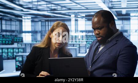 Teamworking colleagues in server farm using laptop to patch network issues affecting transfer rates. Coworkers in data center doing brainstorming, fixing errors affecting supercomputers, camera A Stock Photo