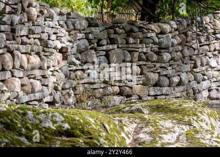 A rustic stone wall made of various sizes of rocks, showcasing a natural, earthy aesthetic. The wall is partially covered with moss, surrounded by gre Stock Photo