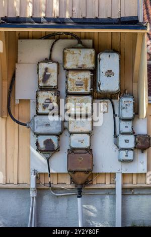 A collection of old, rusty electrical meters and boxes mounted on a wooden wall. The boxes vary in size and shape, showcasing a weathered appearance w Stock Photo