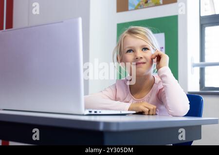 In school, girl sitting at desk using laptop, smiling and thinking Stock Photo