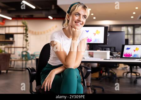 Smiling woman sitting in office, thinking creatively, with computers in background Stock Photo