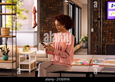 Using smartphone, woman sitting on desk in modern office with sticky notes Stock Photo