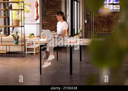 Using laptop, woman sitting on table in modern office, working on project Stock Photo