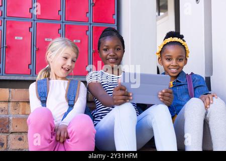 In school, Using tablet, three multiracial girls sitting on steps in front of lockers Stock Photo