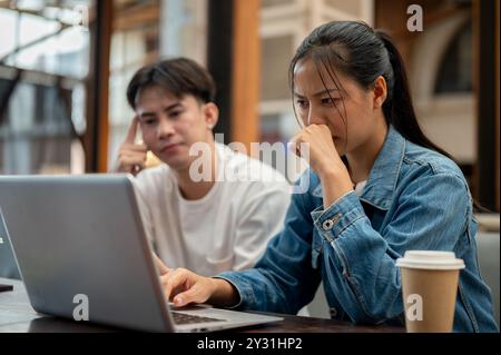 A serious and stressed young Asian woman is working on her laptop while discussing a work issue with her male colleague, looking at her laptop screen Stock Photo