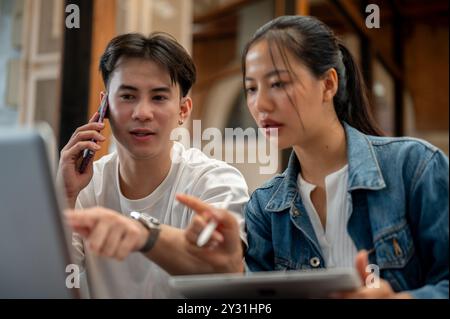 A busy Asian man is multitasking, talking on the phone with someone while discussing work, having a meeting with his female colleague at a coffee shop Stock Photo