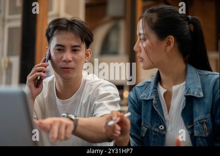 A busy Asian man is multitasking, talking on the phone with someone while discussing work, having a meeting with his female colleague at a coffee shop Stock Photo