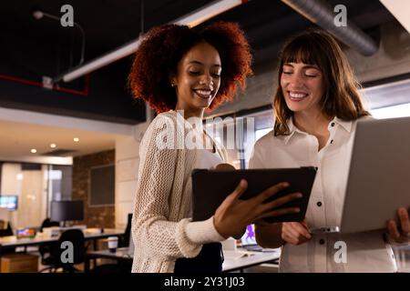Collaborating on project, two multiracial women using tablet and laptop in modern office Stock Photo