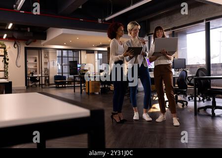 In modern office, three multiracial women collaborating on project using laptop and tablet Stock Photo