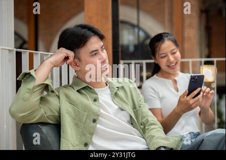An unhappy, annoyed Asian man is sitting on a couch in a coffee shop with a moody expression, feeling ignored by his girlfriend while she is texting o Stock Photo
