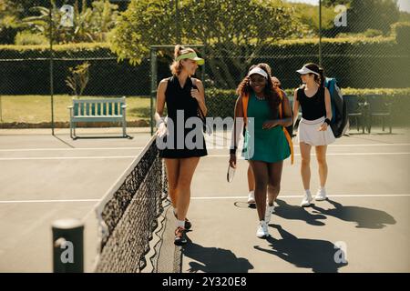 Three women tennis players in sportswear, carrying sports bags, walking together on a court near the net and bench after a game. Stock Photo