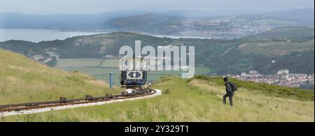 Backpack walker trudging across grassland towards Victorian funicular tram & track climbing up Great Orme views of Llandudno coastline North Wales UK Stock Photo