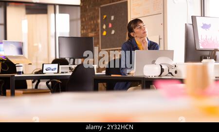 Working in office, woman using laptop and thinking, surrounded by monitors Stock Photo