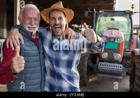 Two excited farmers in hug, father and son, showing thumbs up in front of tractor on cattle ranch Stock Photo