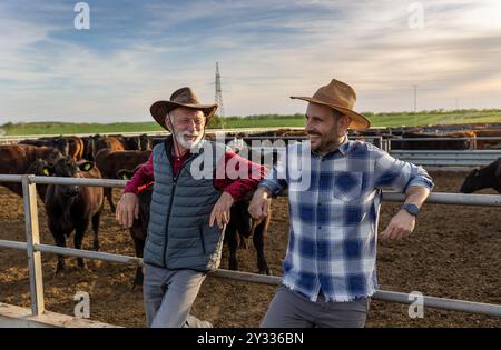 Two satisfied farmers, father and son, leaning on fence of cattle barn and talking on ranch Stock Photo