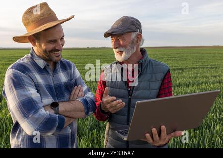 Two smiling farmers standing in green wheat field, using laptop and talking in spring time Stock Photo