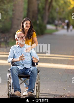 Young woman caregiver pushing senior man in wheelchair in park Stock Photo