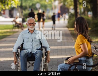 Senior man using wheelchair while young woman sitting on bench and waiting in park Stock Photo