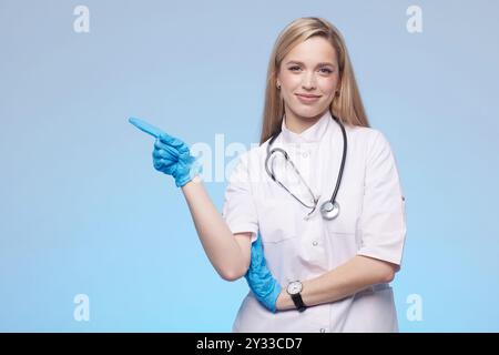 Beautiful lady doctor pointing with hand to copy space, studio shot. Isolated on blue background. Medical student, general practitioner. Stock Photo
